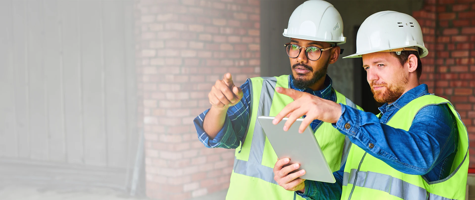 Two construction workers in hard hats and safety vests point while reviewing plans on a digital tablet at a building site.