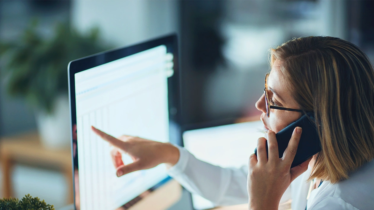 Woman wearing reading glasses is talking on cell phone while pointing to invoices on computer screen