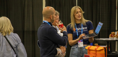 A sponsor at Dimensions demos a solution on her laptop to interested attendees in her booth, wearing company-branded merch.