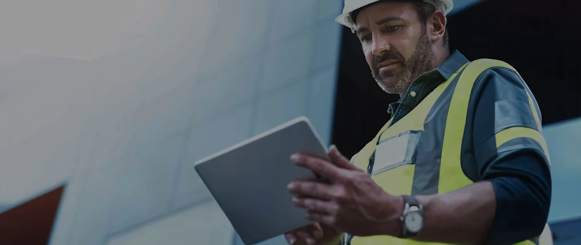 A man wearing a white hard hat and a yellow high-visibility safety vest is shown outdoors, looking intently at a tablet he holds with both hands.