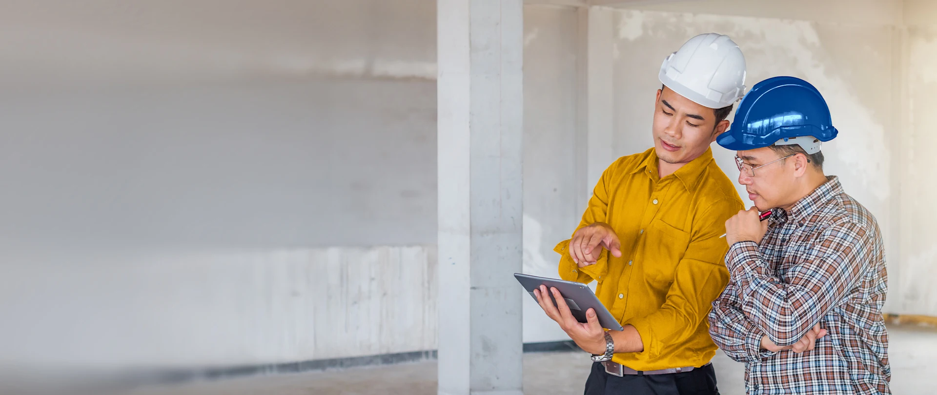 Two engineers in hard hats review project plans on a rugged tablet inside a building under construction.
