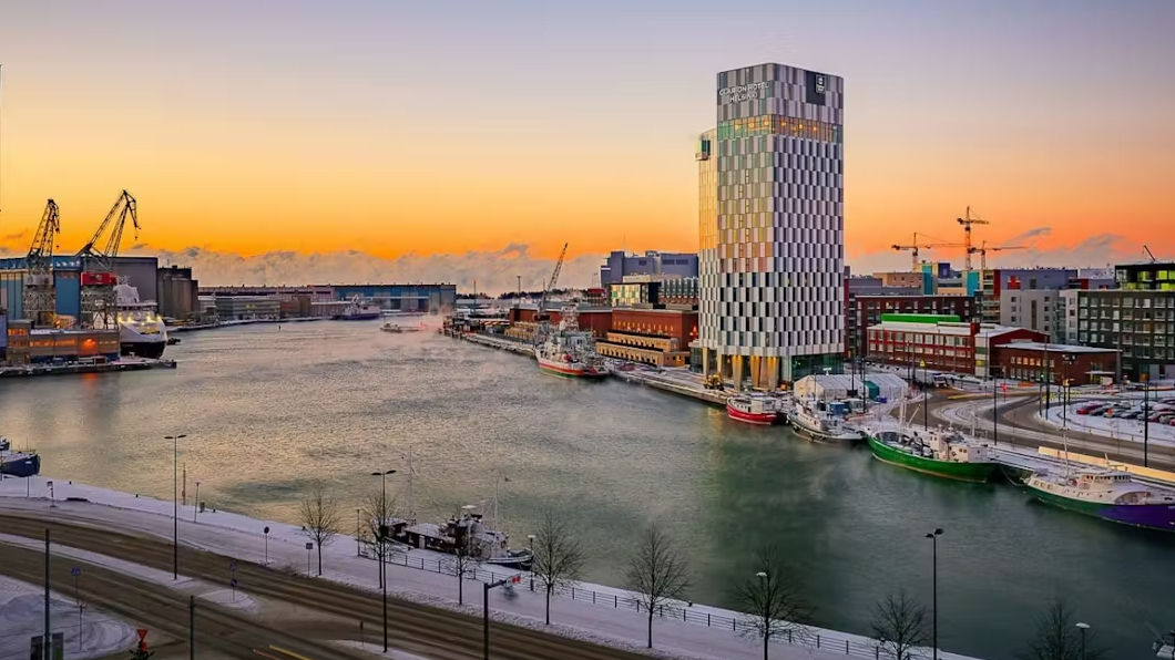 Sunset view of a harbor with a tall, patterned building on the right, ships docked along the snowy waterfront, and cranes in the background.