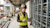 Une femme portant un casque de chantier et un gilet de sécurité sur un chantier utilise une tablette renforcée. Il y a une structure métallique derrière elle.