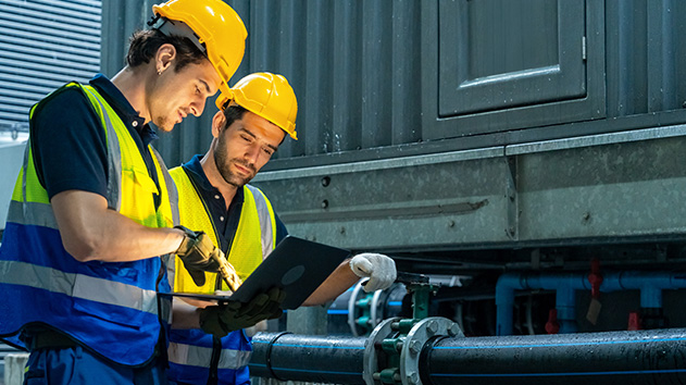 Two HVAC technicians in yellow hard hats and safety vests review data on a laptop while standing next to industrial piping and machinery.