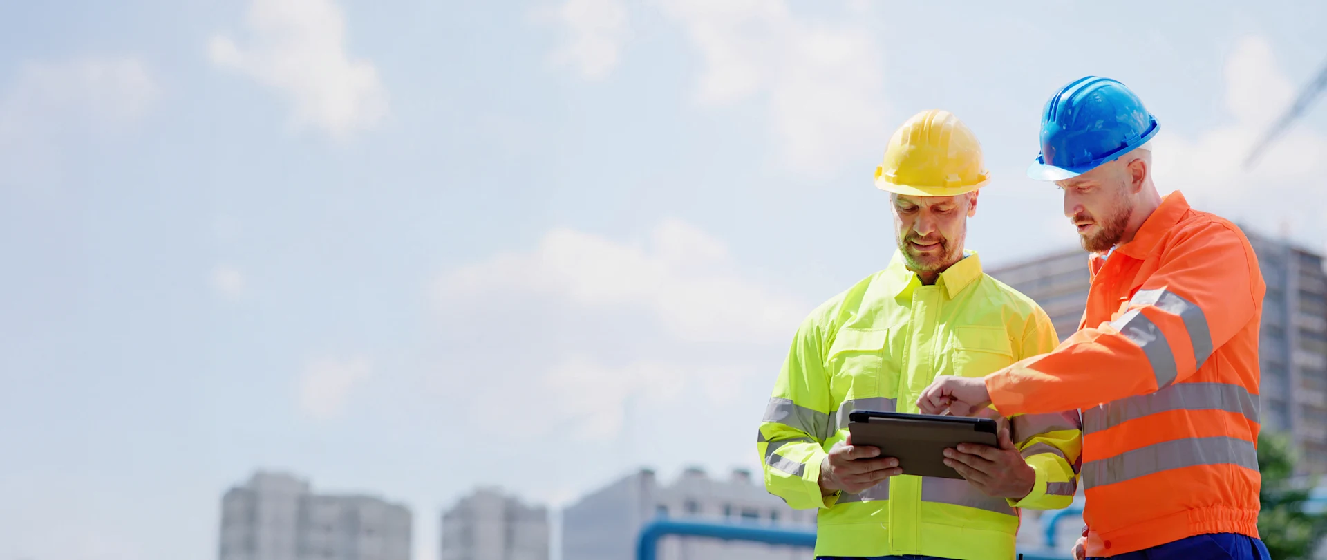 Two construction workers in high-visibility gear and hard hats collaborate on a rugged tablet at an outdoor job site with a city backdrop.