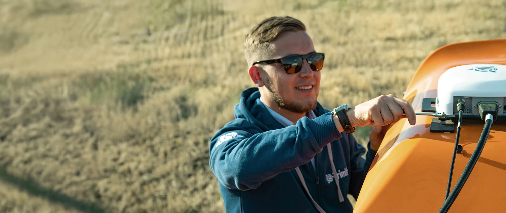 A man wearing sunglasses and a blue hoodie adjusts a white GPS receiver mounted on top of an orange agricultural vehicle in a field.
