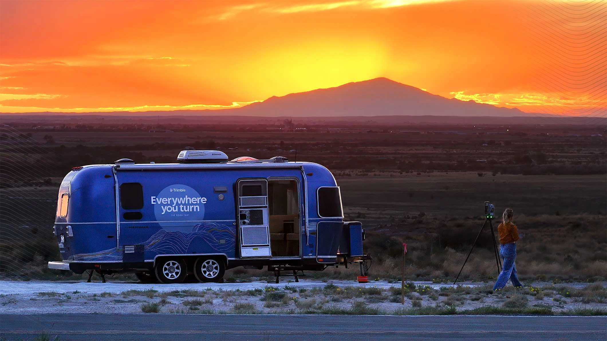 Blauer Trimble Airstream-Trailer im Sonnenuntergang mit dem Logo von „Everywhere You Turn“. Eine Person steht vor einer Bergkulisse neben einem Trimble-X7-Scanner.