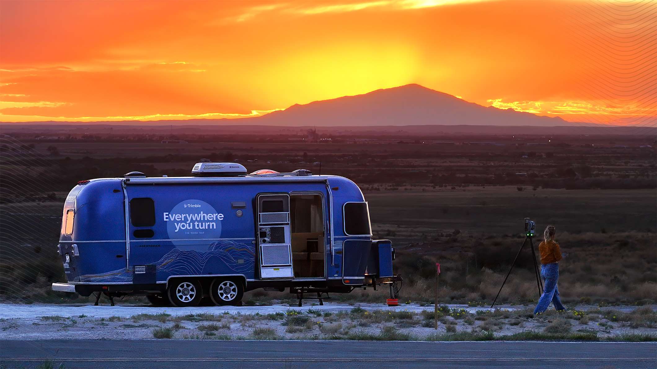 Blue Trimble Airstream trailer at sunset with 'Everywhere you turn' logo. A person stands near a Trimble X7 scanner with a mountain background.