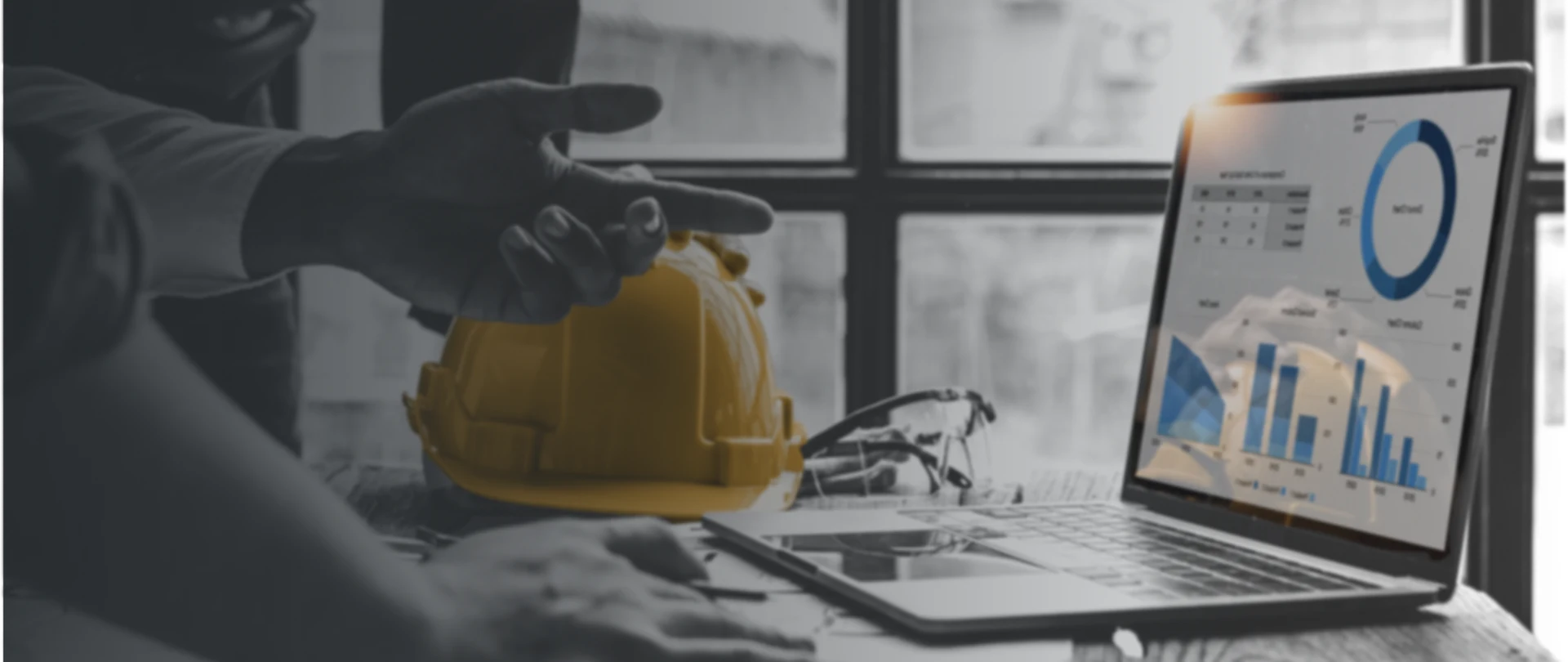 A person gestures near a yellow hard hat while a laptop displays a Trimble construction software dashboard with charts and graphs.