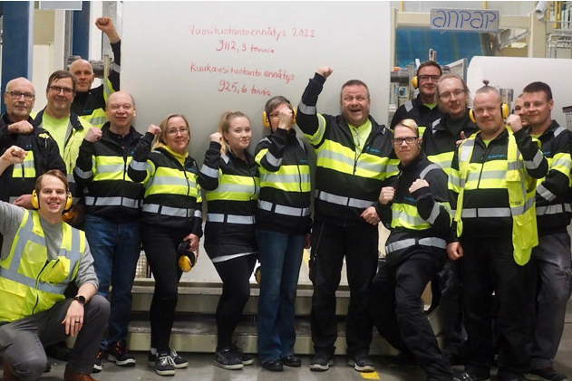 A large group of industrial workers in safety gear celebrate in front of a giant paper roll with Finnish text.