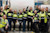 A large group of industrial workers in safety gear celebrate in front of a giant paper roll with Finnish text.