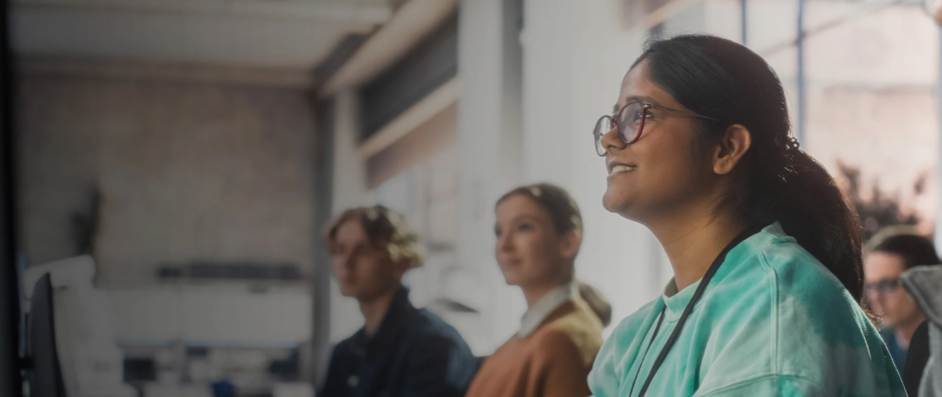 A student wearing glasses smiles while looking forward in a classroom setting with other students.