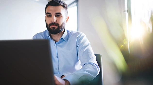 A man with a beard sits at a desk, intently looking at a laptop computer.