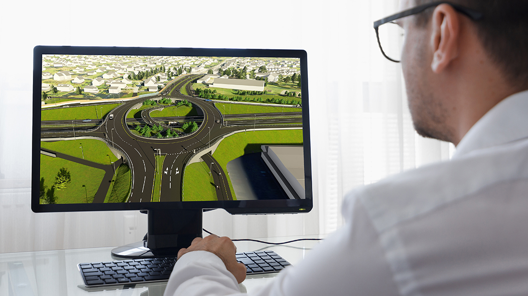 Over-the-shoulder view of an engineer using a computer to design a complex 3D BIM model of a highway interchange and roundabout.