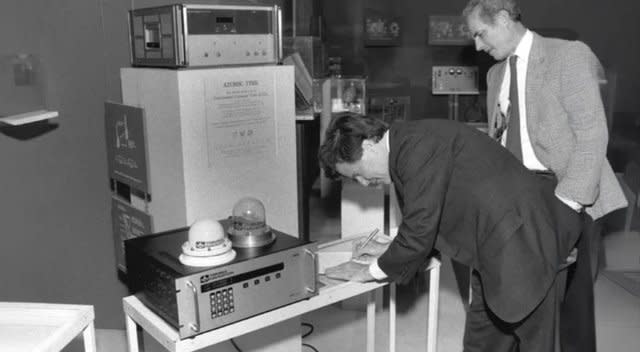 Ein Präzisionszähler von Hewlett-Packard mit GPS-Antennen und einem Schild mit der Aufschrift „Atomic Time“ wird von mehreren Männern begutachtet. Dieses Setup aus den 1980er Jahren war für die Genauigkeit der AECO-Vermessung von entscheidender Bedeutung.