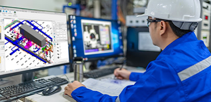 An engineer in a hard hat uses Trimble Nova software to design a complex 3D MEP model on dual monitors in an industrial setting.