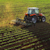 An aerial, slightly angled shot captures an orange tractor with a tiller or cultivator implement working in a field of young, green crops planted in neat rows.