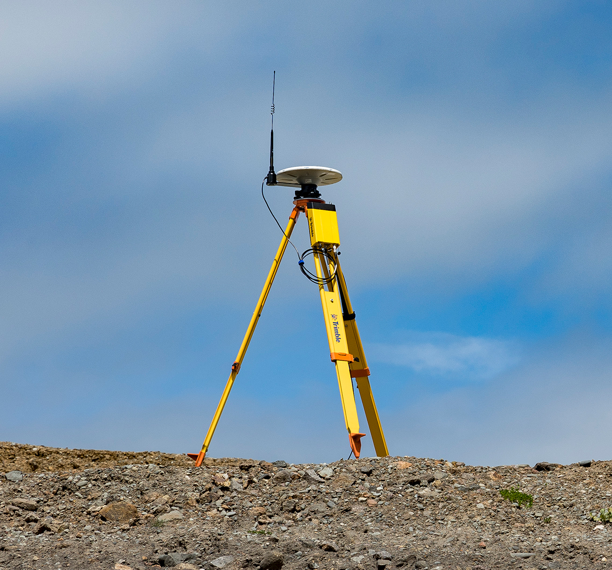 Survey tripod with mounted antenna at the top and the Trimble R750 Model 2 GNSS modular receiver attached below it on a dirt construction site with blue sky and clouds behind it.