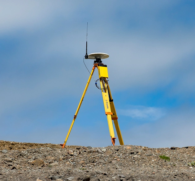 Survey tripod with mounted antenna at the top and the Trimble R750 Model 2 GNSS modular receiver attached below it on a dirt construction site with blue sky and clouds behind it.