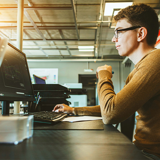Man with yellow sweater at desk looking at computer showing graphs