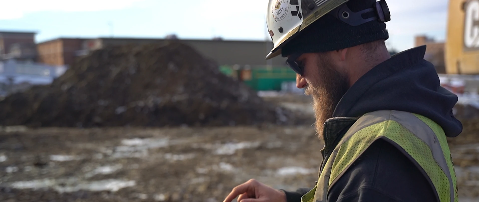 A construction worker uses a Trimble rugged tablet on a job site. He wears a hard hat with a Portland logo and a high-visibility safety vest.