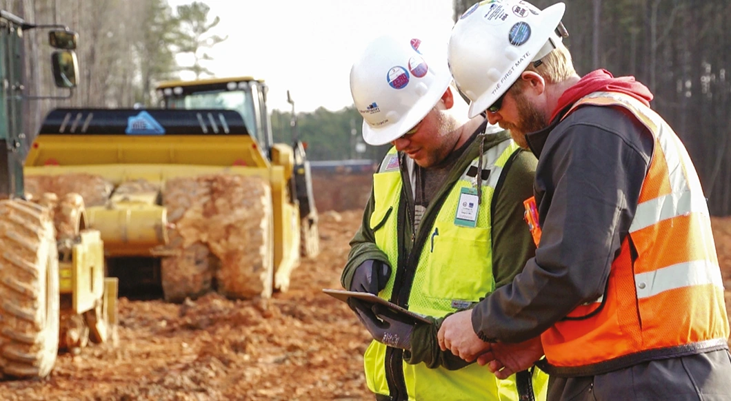 Two construction workers on a Mortenson site review a tablet next to heavy machinery featuring Trimble Earthworks technology.