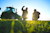Two agricultural workers, silhouetted against a bright sunset, inspect young crops in a field, with one gesturing, and a large green tractor partially visible behind them.