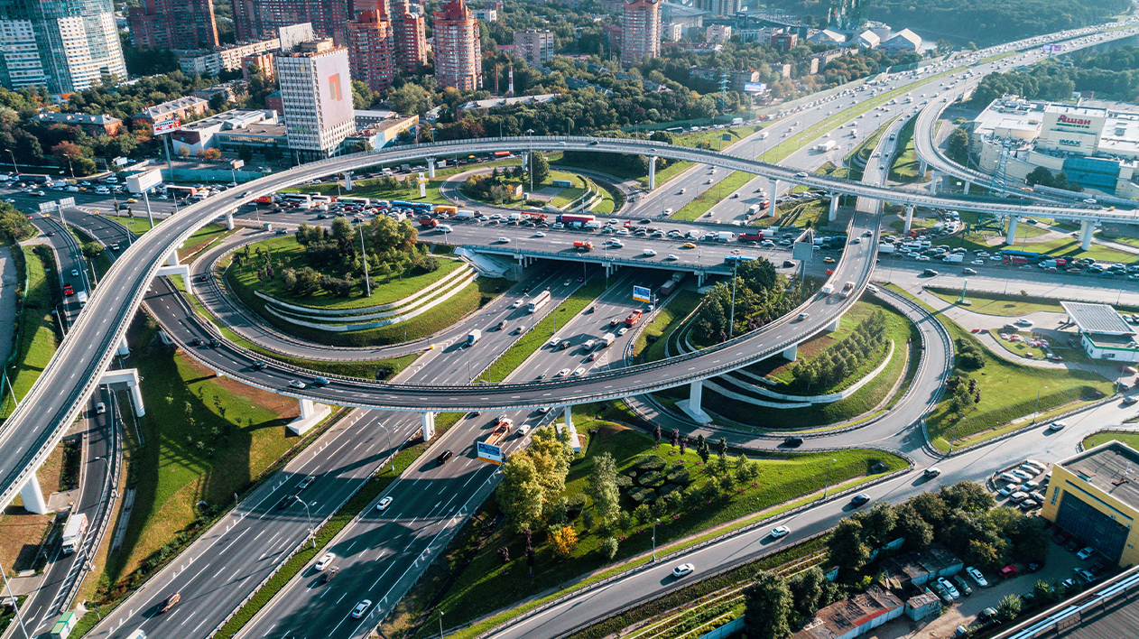 An aerial shot captures a massive, complex highway interchange and overpass system in a modern city on a sunny day.
