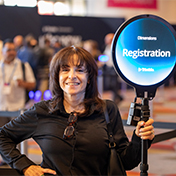 A smiling woman stands at a Trimble Dimensions conference holding a glowing blue circular sign that reads "Dimensions Registration" with the Trimble logo.