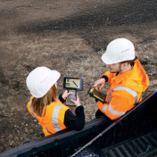 Two construction workers in safety gear review technical data on a tablet at an outdoor work site.