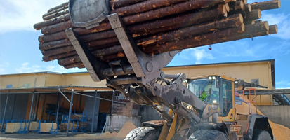 A front loader lifts a stack of logs in an industrial yard, with a yellow building in the background under a bright blue sky