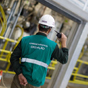 A worker in a green vest and white hard hat, labeled "OPERAÇÃO," communicates via radio in an industrial setting with yellow railings and machinery.