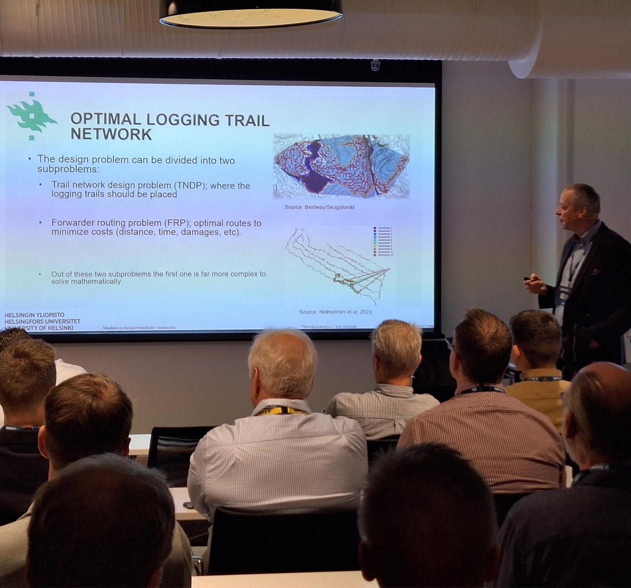 A lecturer presents on "Optimal Logging Trail Network" in a classroom. The slide shows text and maps. Audience members are seated attentively.