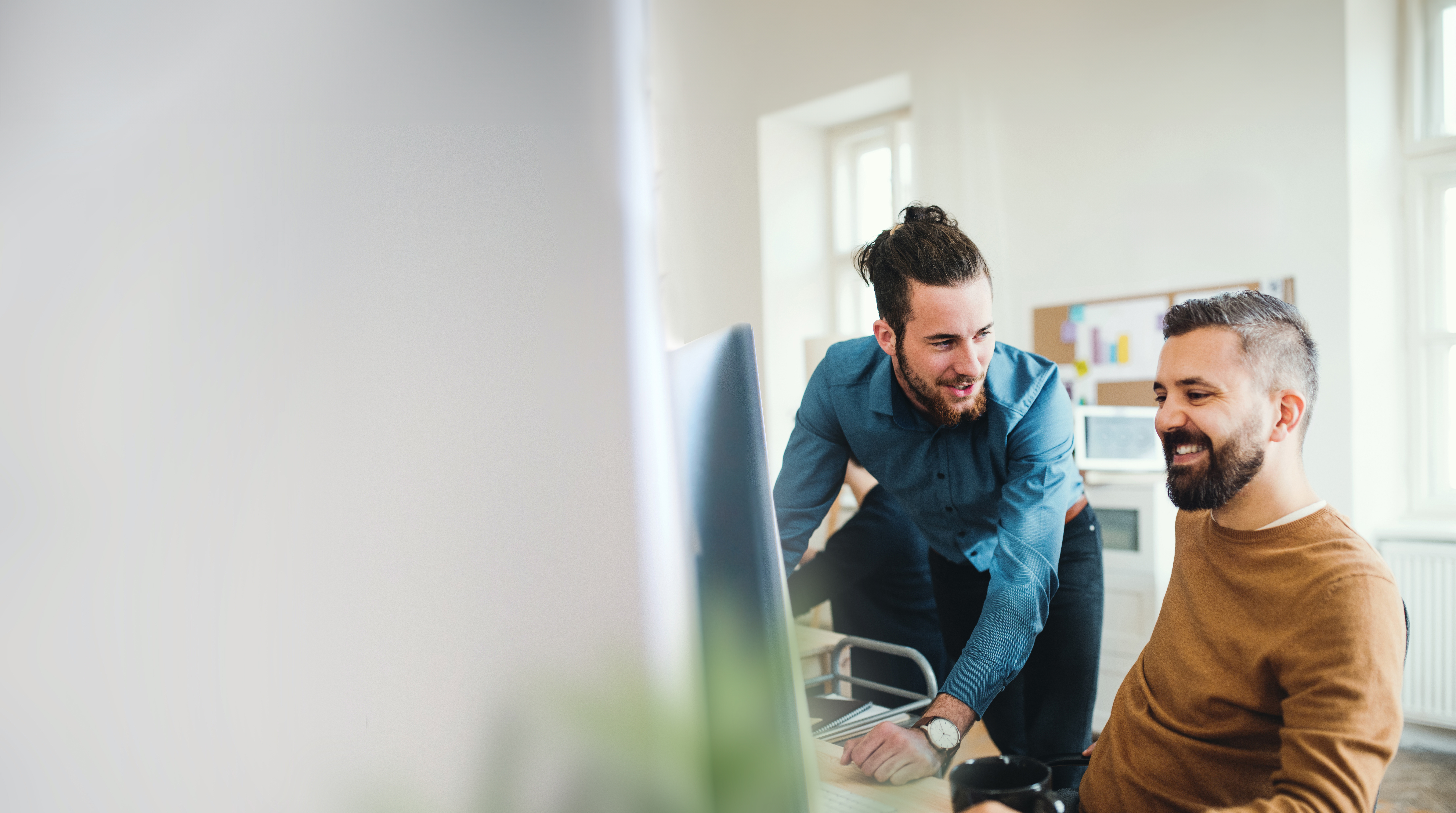 Two office professionals collaborate at a desk in a bright, modern workspace. One man sits at a computer while the other leans in from the side to look at the screen; both are smiling and engaged in discussion.