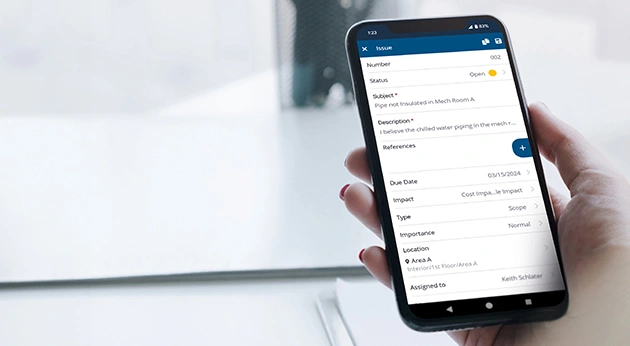 A hand holds a smartphone displaying a construction issue report on the Trimble ProjectSight mobile app.