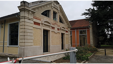 A building with a mix of light-colored and red brick, arched windows, and a prominent gable, with a vehicle barrier in the foreground.