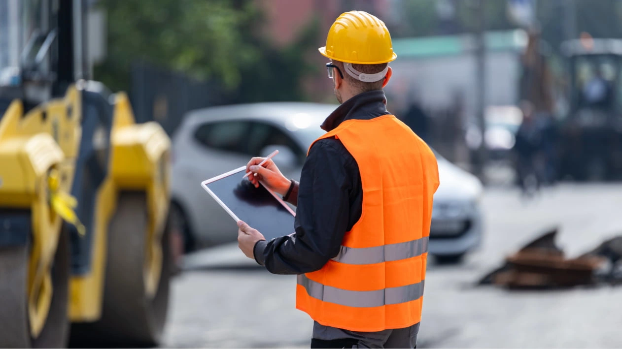 Worker in an orange safety vest and yellow hard hat using a tablet at a busy road construction site.