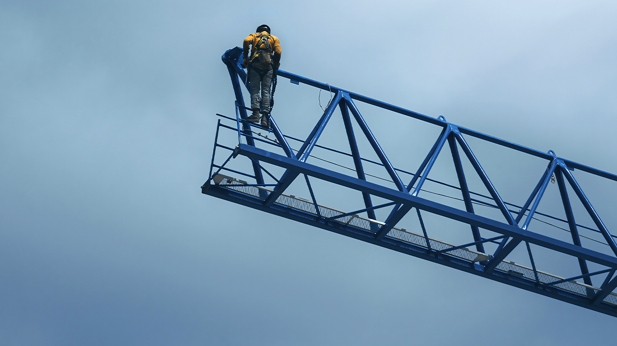A low-angle shot from below captures a person in safety gear, including a yellow jacket and a harness, standing at the end of a long, blue truss or boom.