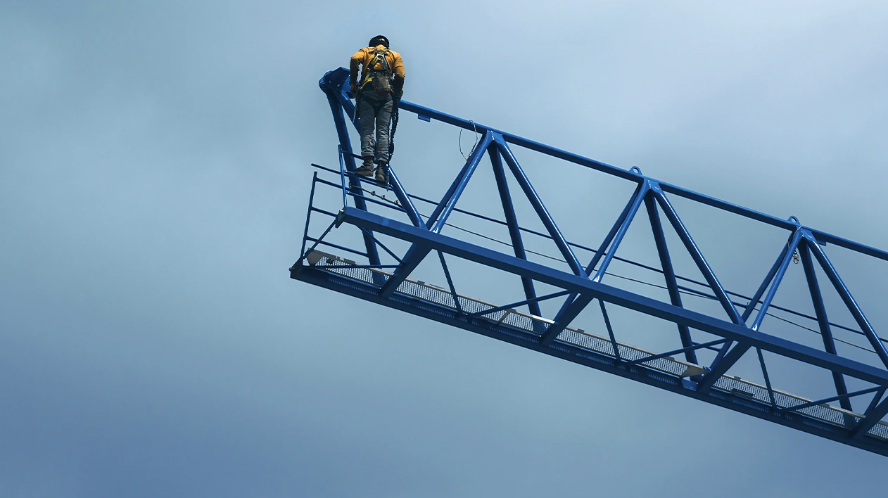 A low-angle shot from below captures a person in safety gear, including a yellow jacket and a harness, standing at the end of a long, blue truss or boom.
