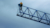 A low-angle shot from below captures a person in safety gear, including a yellow jacket and a harness, standing at the end of a long, blue truss or boom.