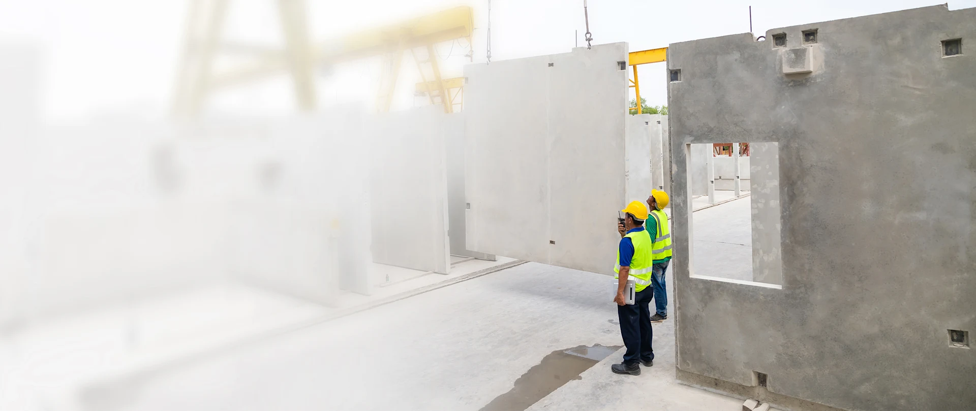 Two construction workers in safety gear at a precast concrete facility monitor a crane lifting a large wall panel.