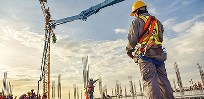 A wide, low-angle shot captures a construction site at dusk or dawn, with a worker in a yellow hard hat and safety harness standing in the foreground, viewed from behind, looking out over the site.