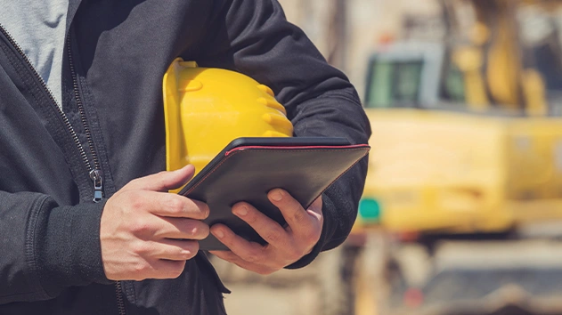 A construction worker holds a yellow hard hat and a tablet with red trim. A blurred yellow excavator is visible in the background.