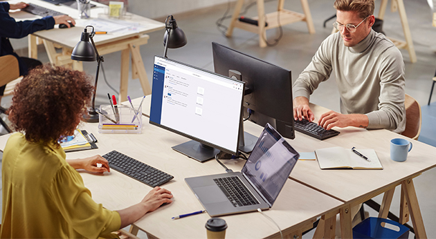 Two professionals work in a modern office environment using Trimble Connect software and data analytics dashboards on their computers.