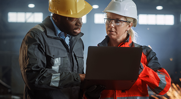 Two industrial workers in hard hats and safety gear collaborate on a laptop in a factory setting with sparks in the background.
