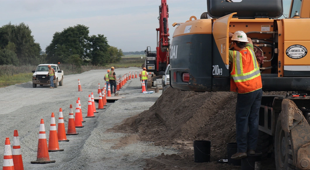 Worker services a Hyundai excavator on a road site lined with orange cones, illustrating Trimble B2W Maintain maintenance software.