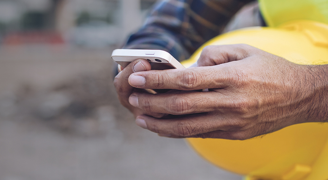 A close-up of a construction worker in a plaid shirt holding a white smartphone, with a yellow hard hat tucked under their arm at a job site.