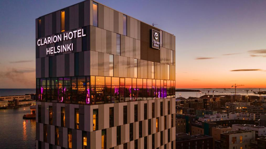 Modern hotel building with large windows and "Clarion Hotel Helsinki" sign, overlooks a harbor cityscape at sunset.
