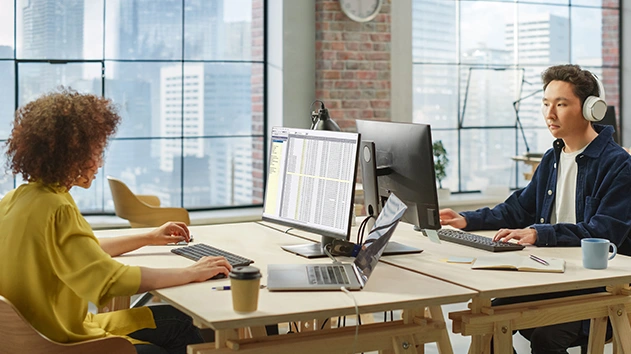 Two people working at a desk with computers in a modern office space with large windows.