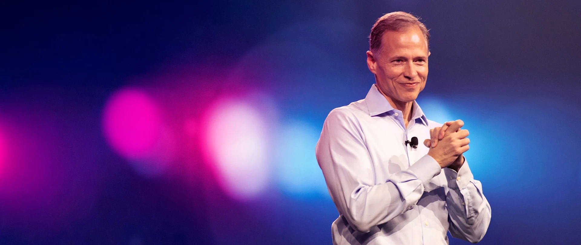 Rob Painter, Trimble President and CEO, smiling on stage during a presentation with pink and blue bokeh lights in the background.